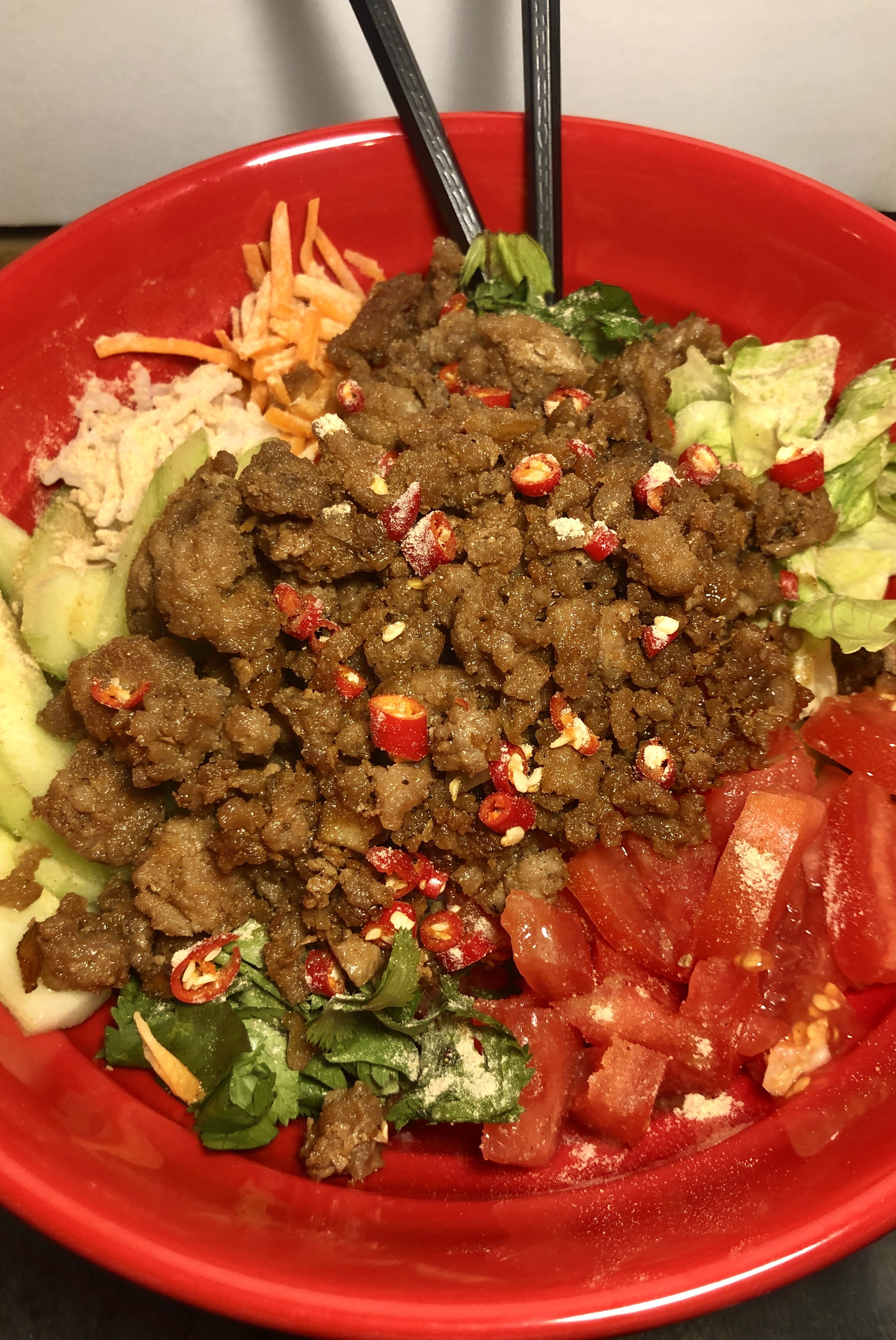 Thai ground pork rice bowl close up shot. The rice bowl has vegetables covering the rice along the edge of the bowl. The caramelized ground pork is in the middle of the bowl with toasted rice powder and Thai chilis over the top.