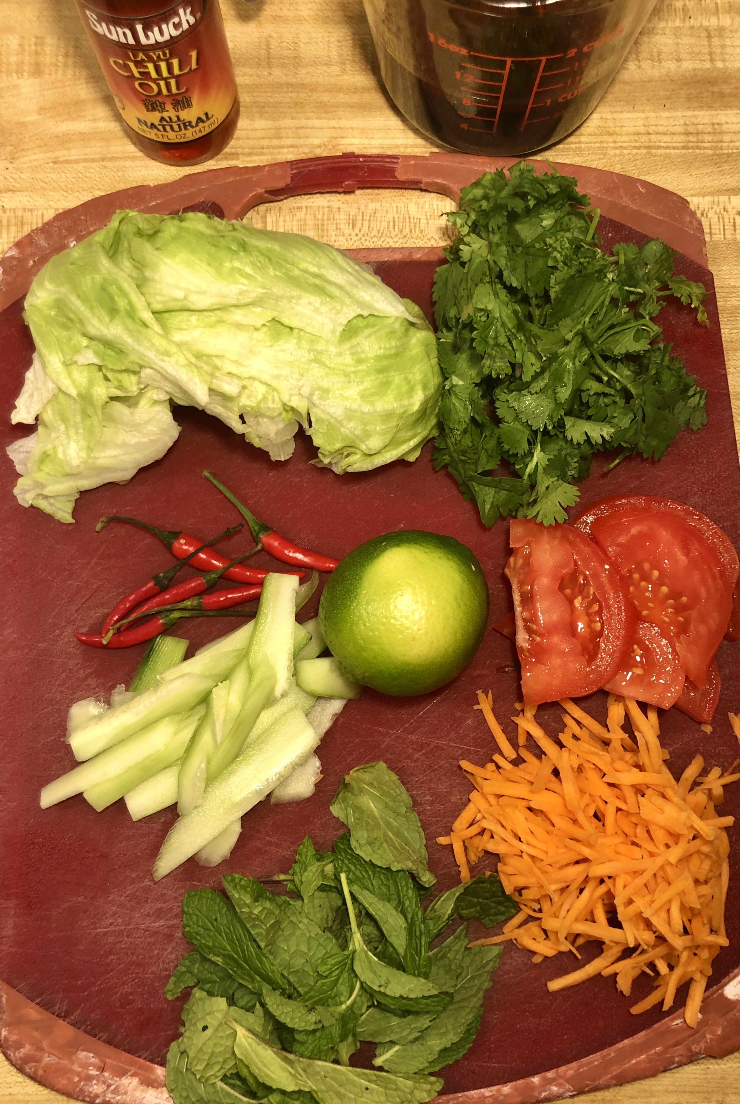 Red cutting board with the vegetables to make the rice bowl on top. These include lettuce, cilantro, Thai chilis, lime, cucumber, tomato, carrots and mint. Chili oil and the sauce for the recipe are pictured in the background.