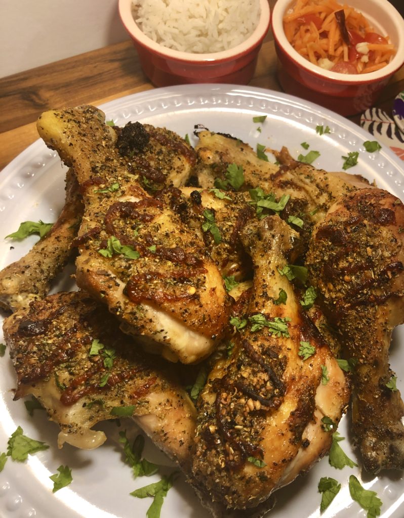 Baked honey sriracha chicken drumsticks plated on a white plate and garnished with chopped cilantro. The plate is on a wooden table with two ramekins in the background. One ramekin has Jasmine rice and one has som tum (Thai carrot salad).