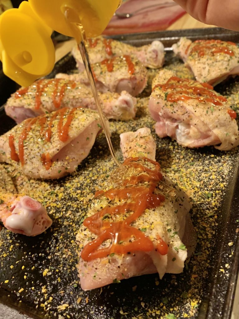 Chicken drumsticks lying on a baking dish and seasoned with garlic powder, Chicago steak seasoning, lemon pepper and other seasonings. Sriracha is covering the seasonings and honey is being drizzled from a bottle over the top of a drumstick.