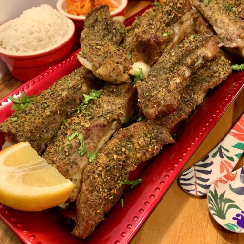 Seasoned oven baked pork ribs stacked and plated on rectangle serving dish with a wedge of lemon. There are two red ramikens in the background. One with Jasmine rice and one with som tum. Floral utensils are pictured beside the serving dish.