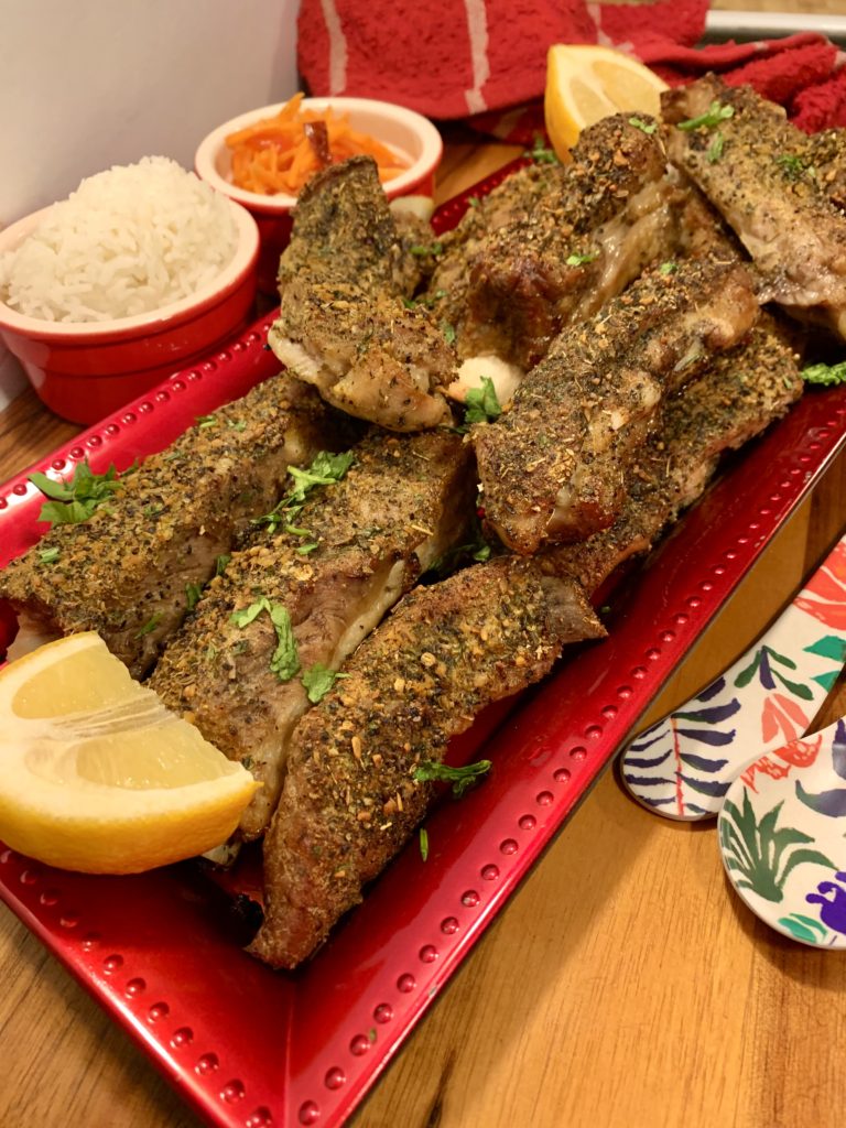Seasoned oven baked pork ribs stacked and plated on rectangle serving dish with a wedge of lemon. There are two red ramikens in the background. One with Jasmine rice and one with som tum. Floral utensils are pictured beside the serving dish.