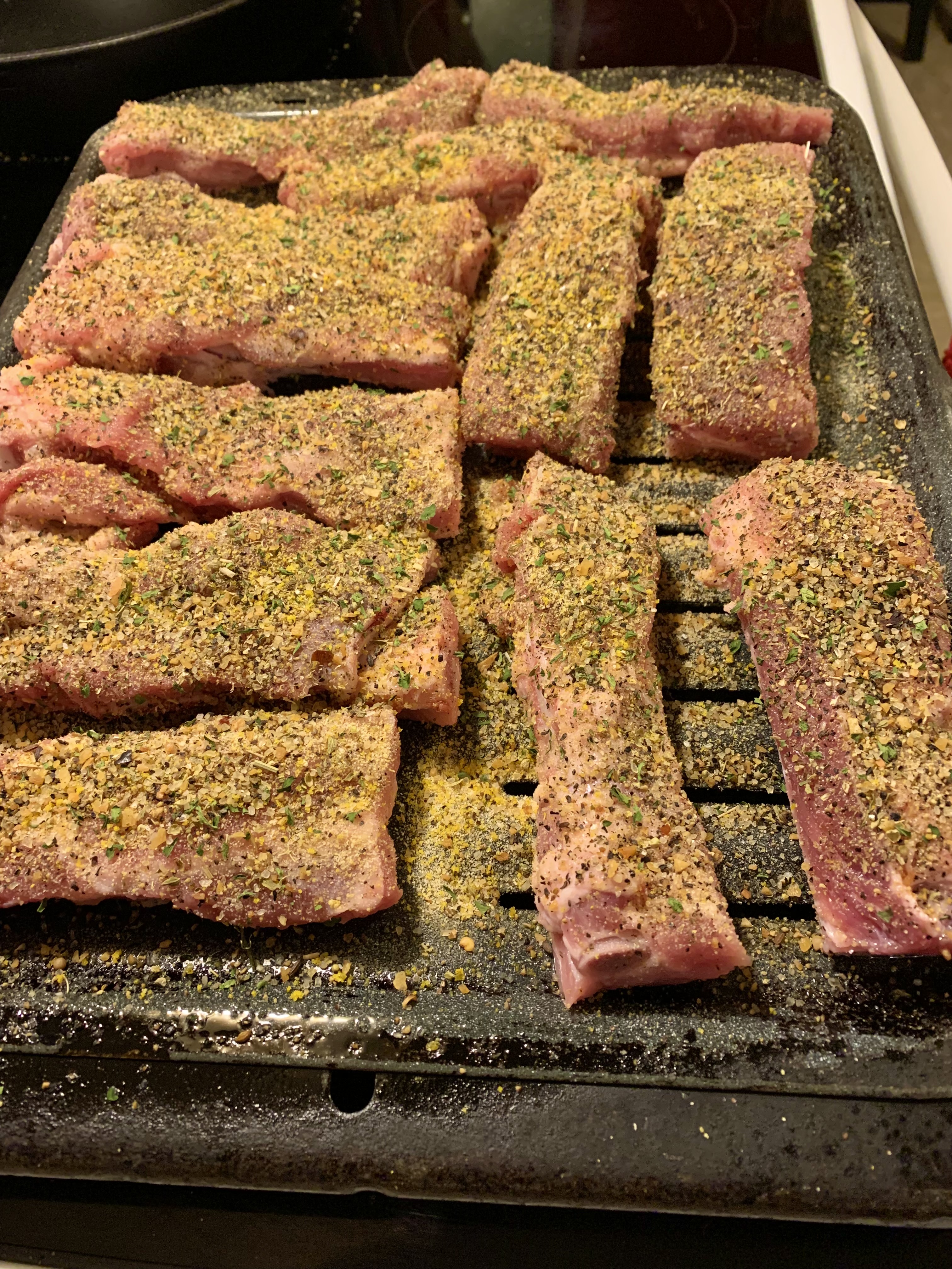 Sliced pork ribs are seasoned and ready to go into the oven. They are pictured on a broiling pan.