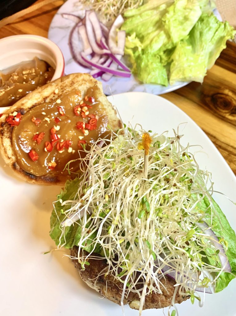Thai pork burger on white square plate and topped with bok choy, sprouts and purple onion. The top bun is beside the burger on the plate and topped with peanut sauce and thinly sliced Thai chili peppers. There is a red ramiken with additional peanut sauce behind the plate. There is a white marble plate with additional vegetables for topping. Everything is pictured on a wooden cutting board.