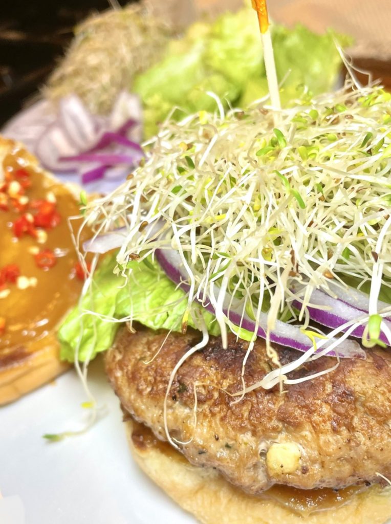 Thai pork burger close up showing the bok choy, sprouts and purple onion toppings. The burger is pictured next to the top bun covered in peanut sauce in thinly sliced Thai chili peppers. There is a plate in the background with additional veggies.