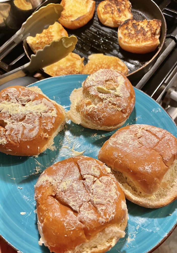Blue plate with hamburger buns that have been covered in butter. In the background is a grill pan with hamburger buns toasting.