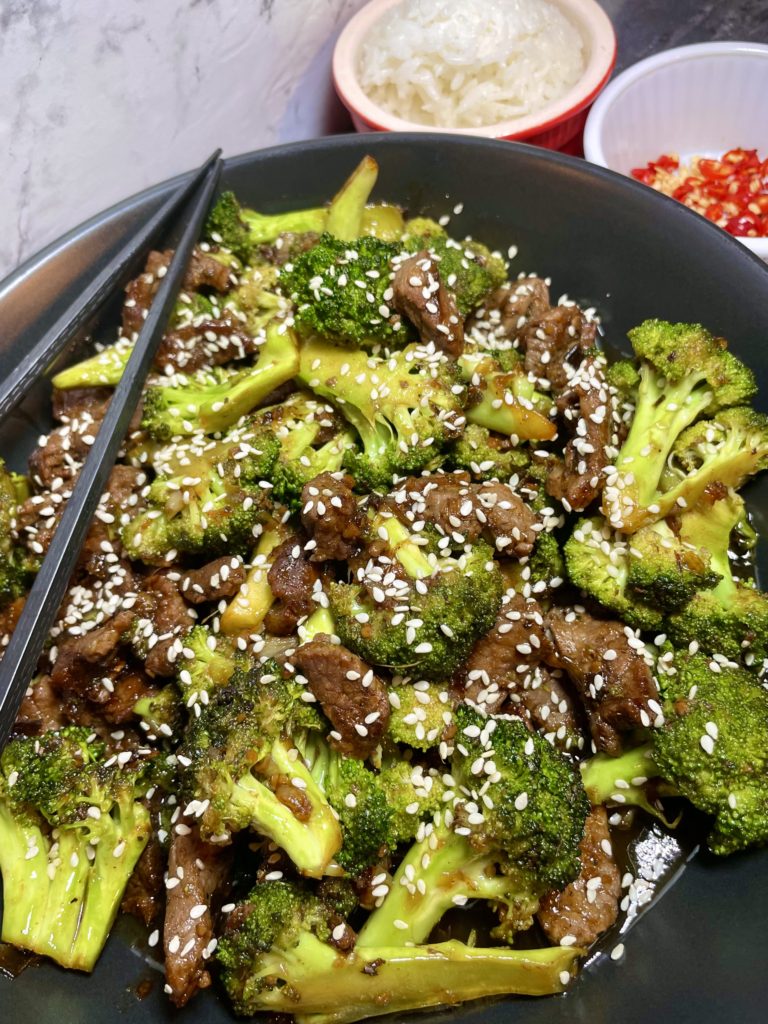 Better Than The Buffet Beef and Broccoli pictured in a black bowl with black chopsticks across the top of the bowl. The dish is garnished with sesame seeds. There are ramekins in the background. One has Jasmine rice and one has fresh Thai chili peppers that have been sliced.