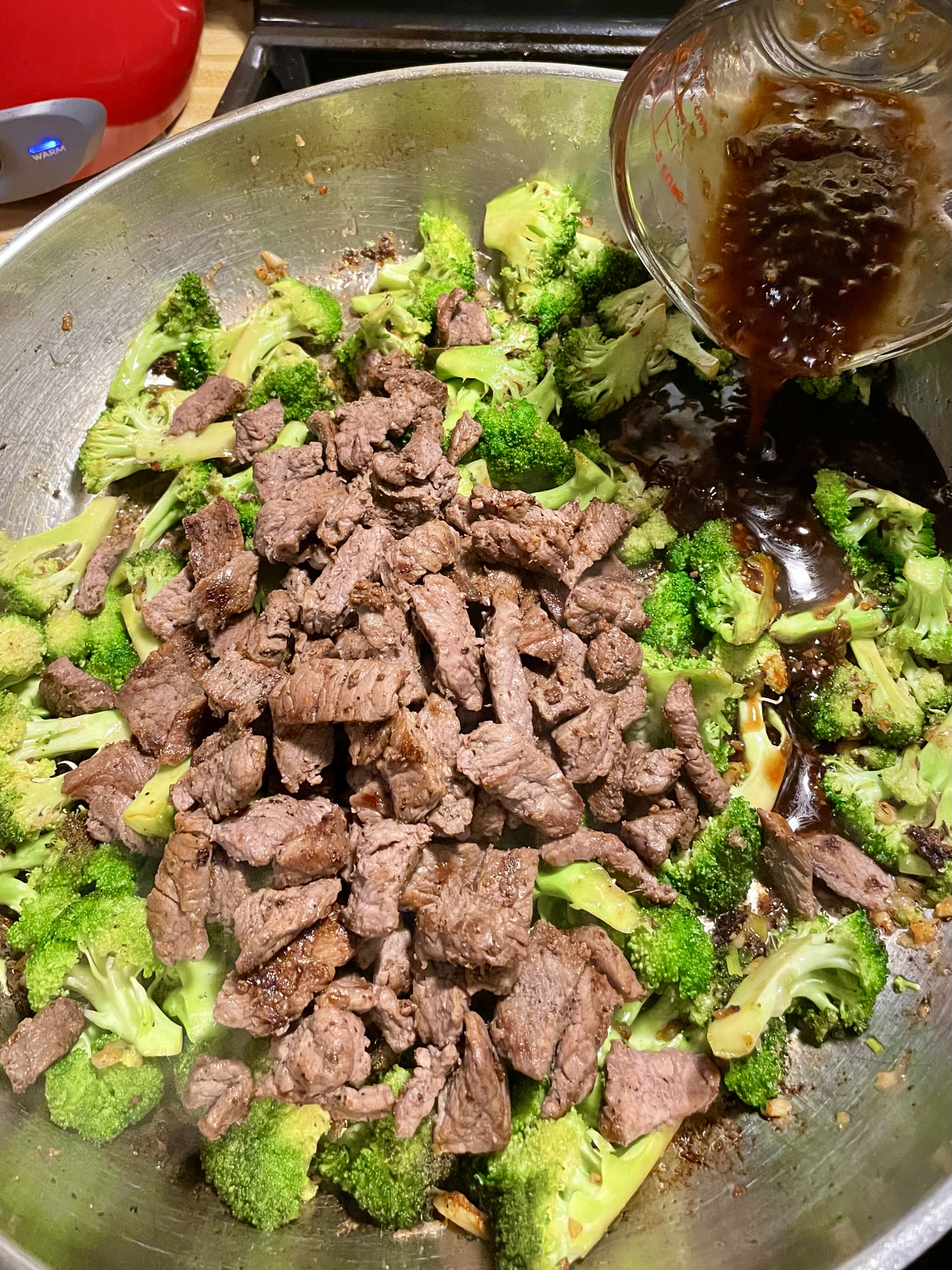 Garlic and broccoli frying in a large skillet. Thinly sliced cooked angus steak and beef and broccoli sauce are being added to the pan.
