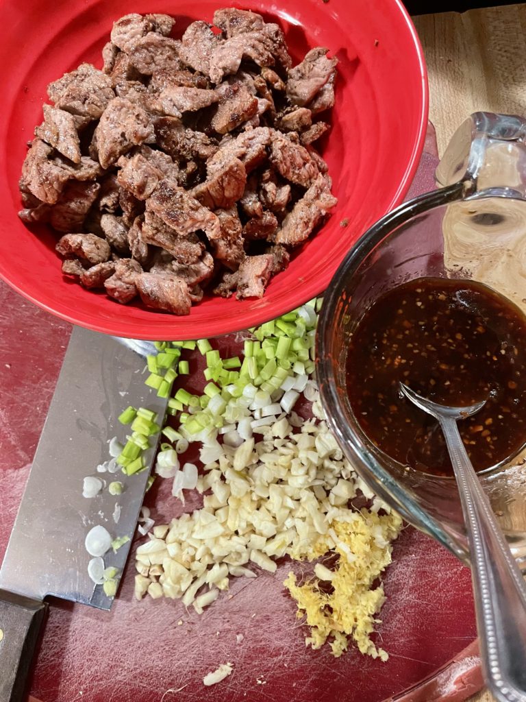 Cooked angus steak is pictured in a red bowl on a red cutting board. There is chopped garlic, green onion and grated ginger on the cutting board beside a knife. The beef and broccoli sauce is on the cutting board mixed together in a glass measuring cup.
