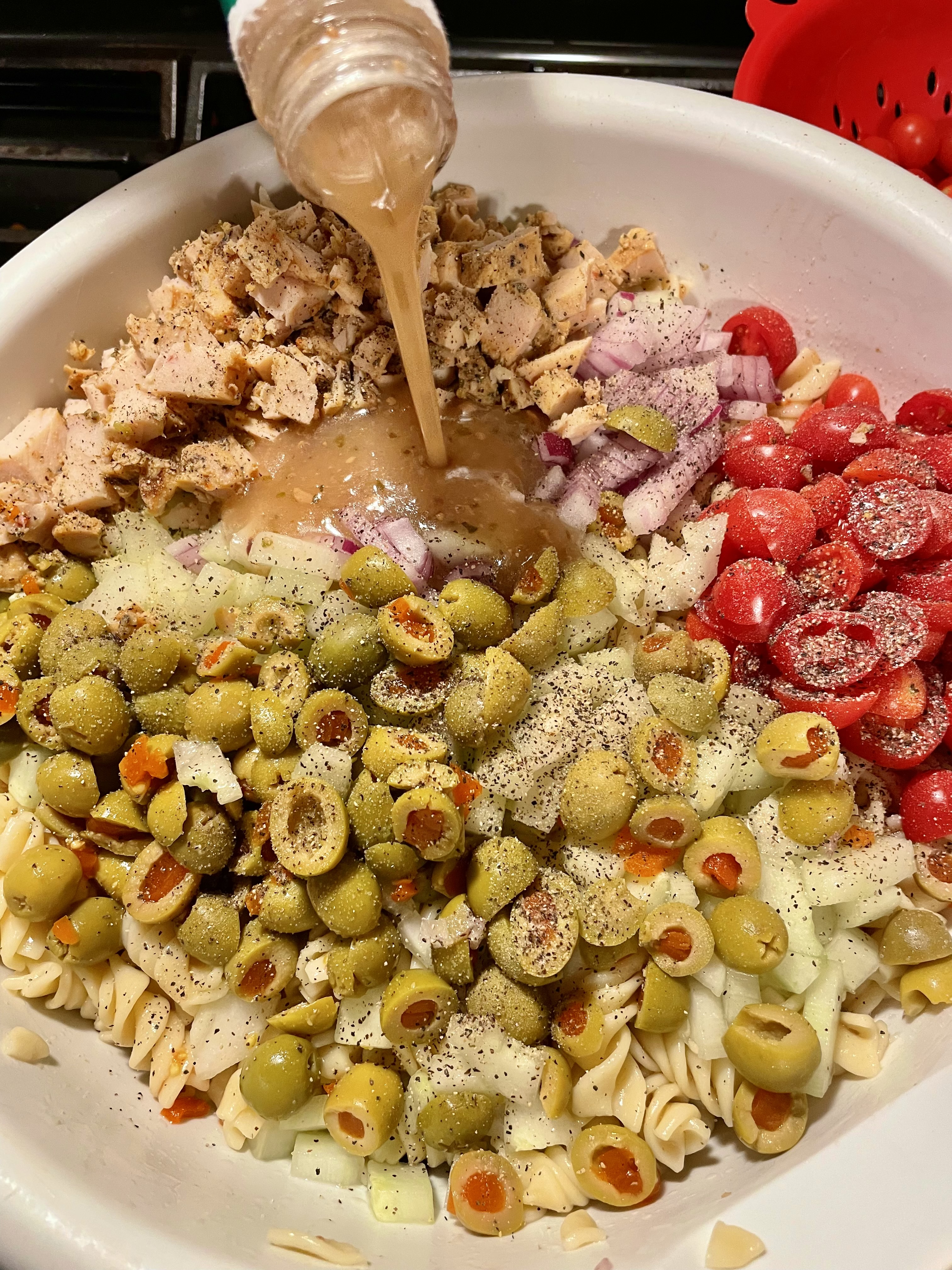 Rotini, cherry tomatoes, cucumber, green olives and grilled chicken in white mixing bowl. Greek dressing is being poured into the bowl.