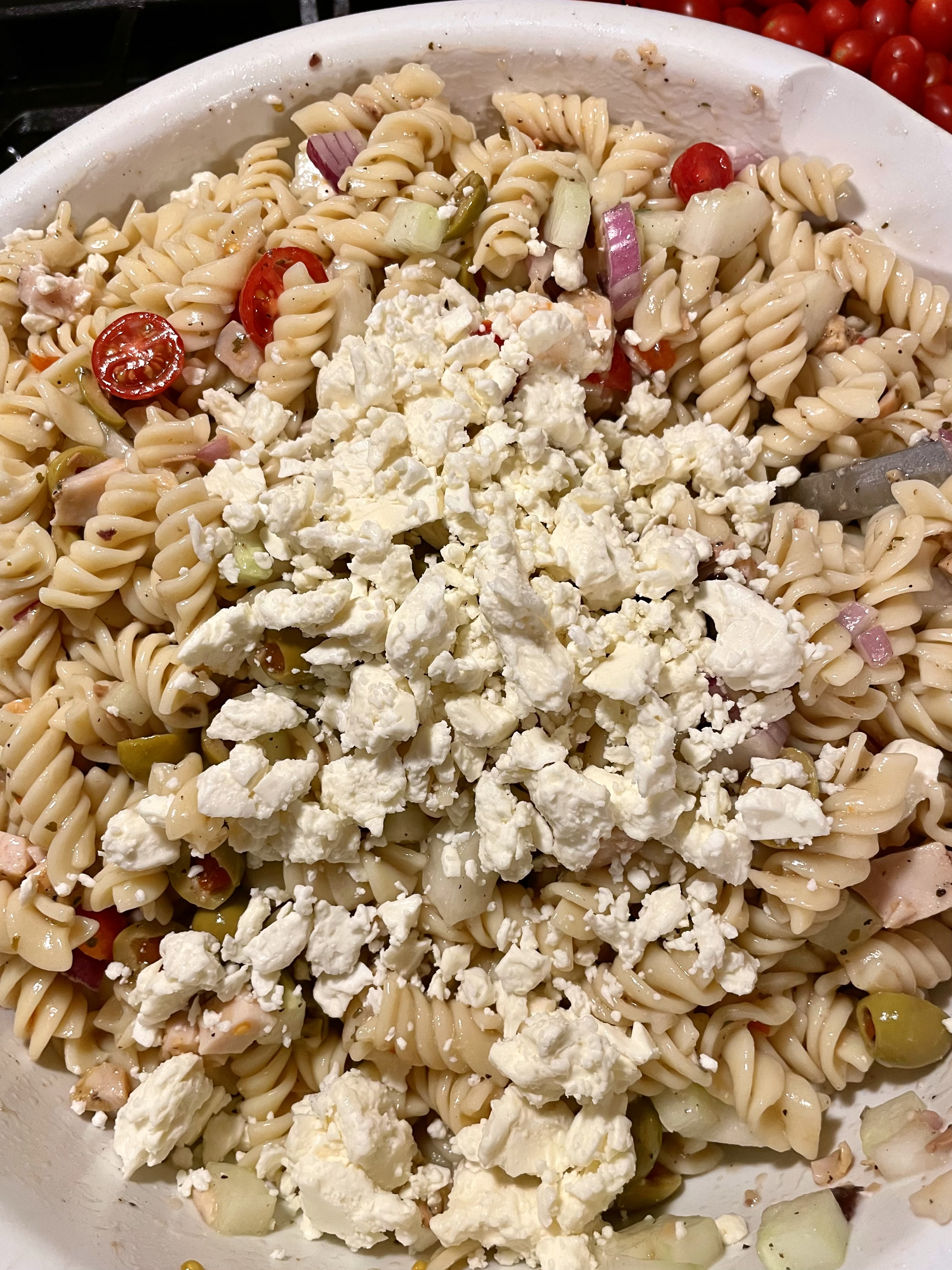 Mixing bowl with rotini, tomatoes, cucumber, olives and onion with feta being added in.