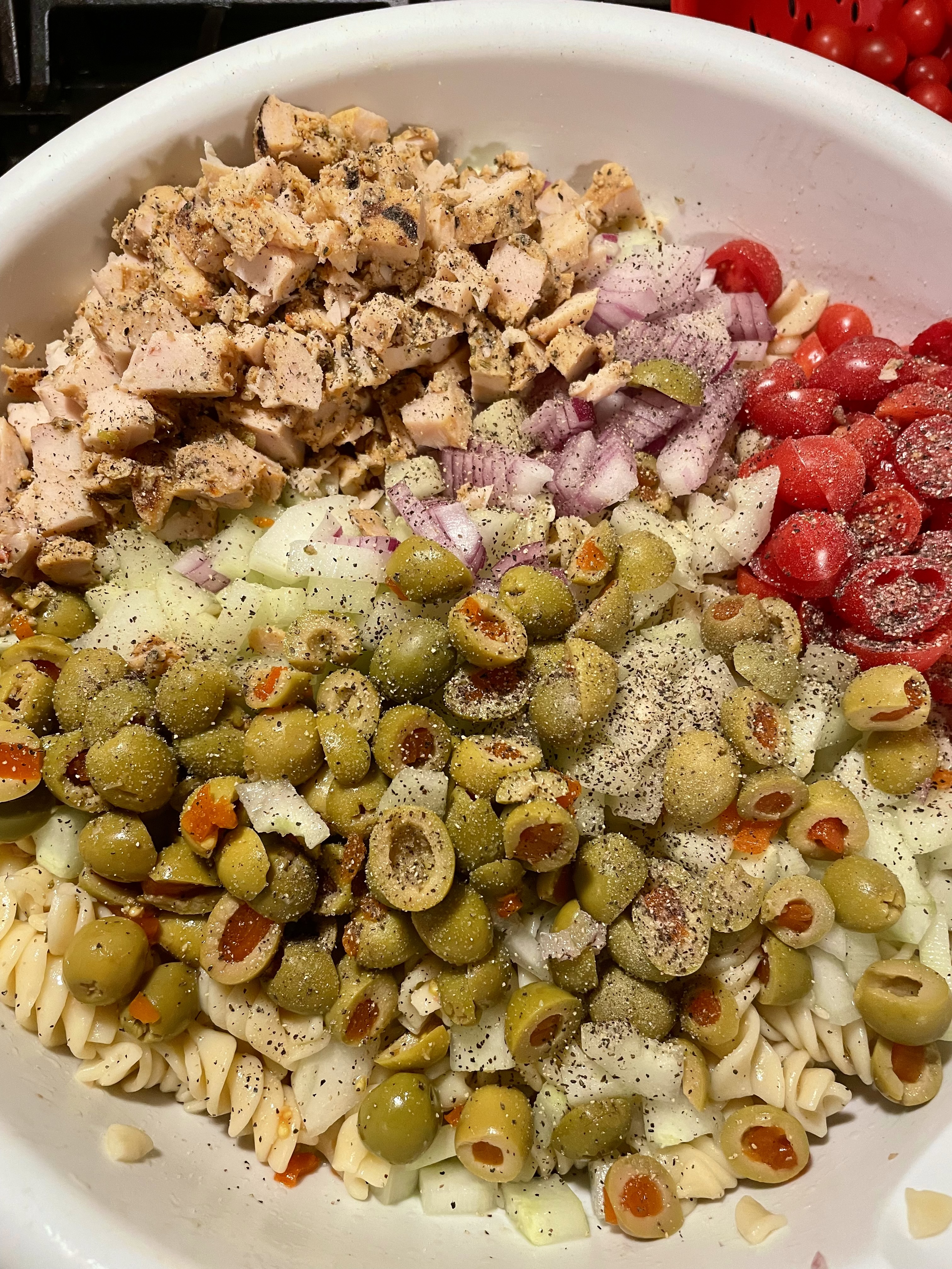 White mixing bowl with rotini, chicken, red onion, cherry tomatoes and green olives.