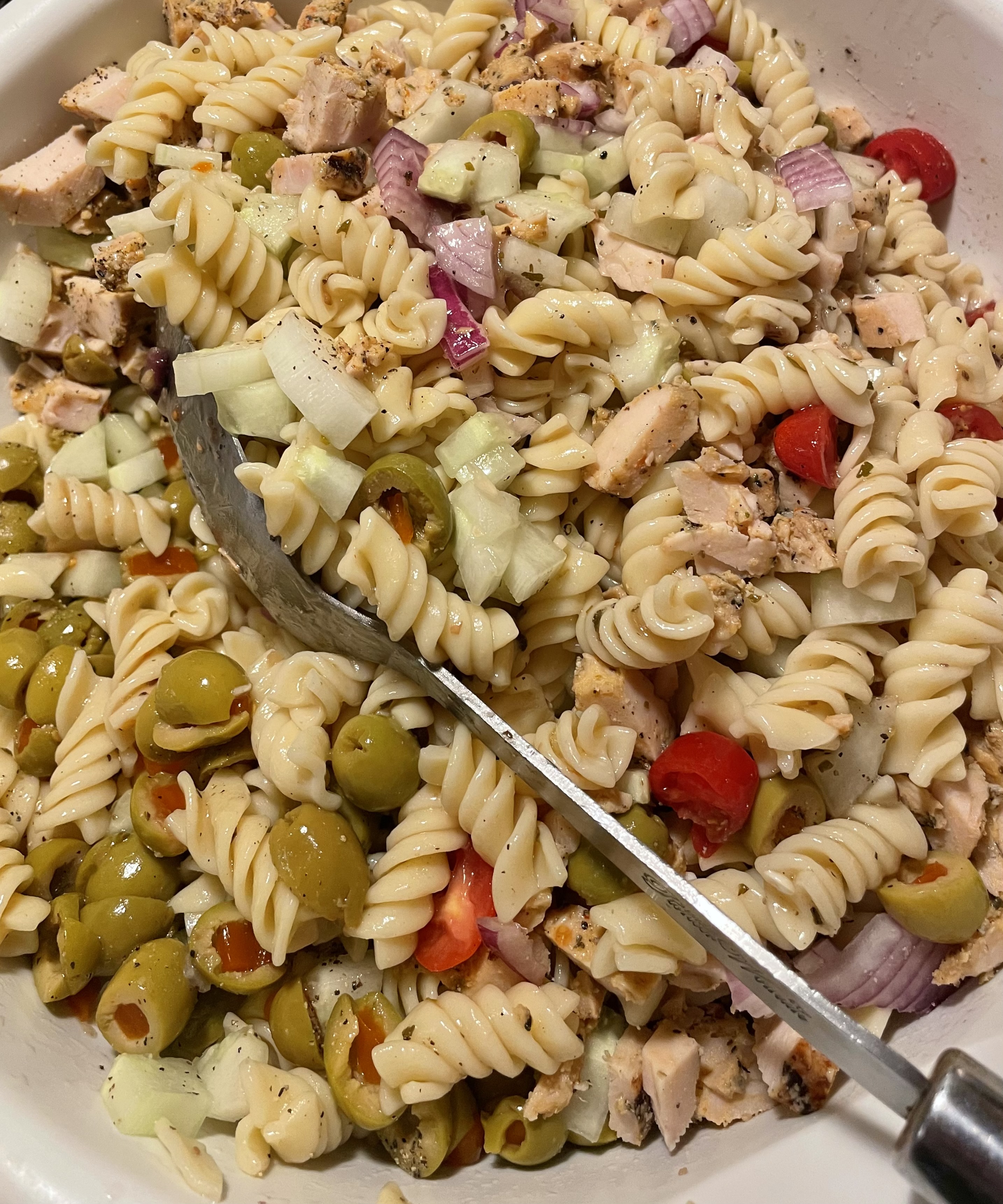 Greek pasta salad being mixed together with a silver mixing spoon in a large white mixing bowl.