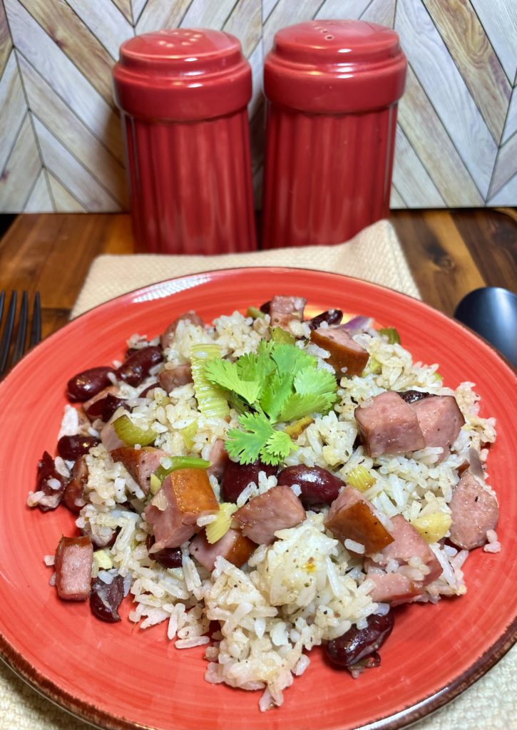 Red Beans and Rice pictured with a cilantro garnish on top. The red beans and rice is on a red plate over a tan cloth on a wooden cutting board. There are oversized red salt and pepper shakers in the background and matte black fork and spoon beside the red plate.