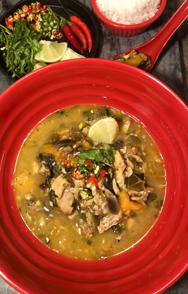Moqueca (Brazilian Fish Stew) plated in red bowl and garnished with cilantro and fresh Thai chilis. There is a mini cast iron skillet in the background with lime wedges, cilantro and Thai chili peppers. There is a red ramekin in the background with white rice. Everything is on top of a dark gray marbled surface.