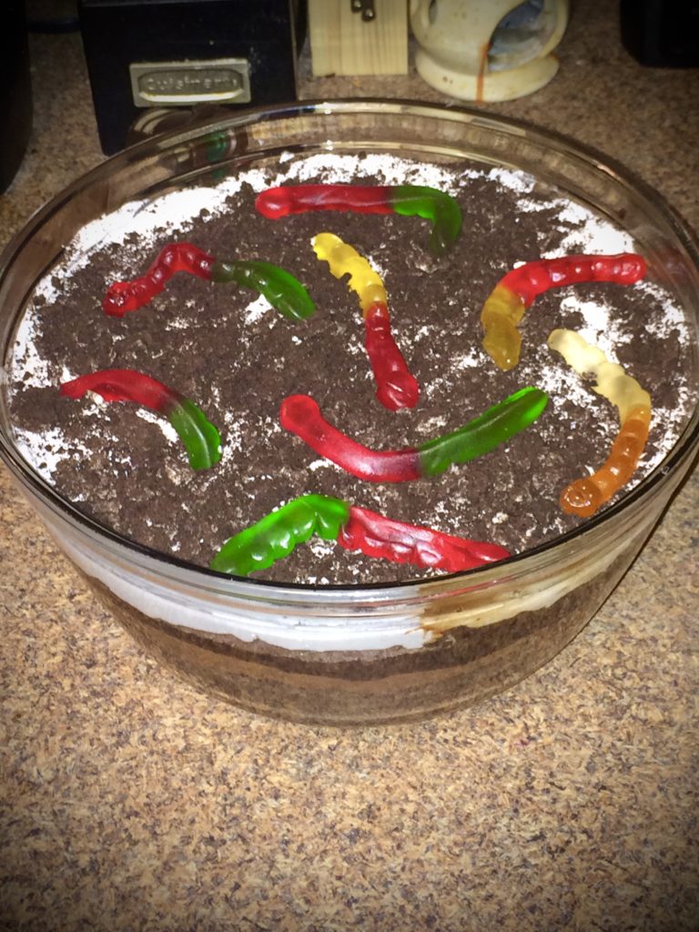 Easy Chocolate Dirt Cake dessert pictured in glass trifle bowl. Layers of chocolate can be seen and the dessert is topped with cool whip, crushed Oreos and gummy worms.