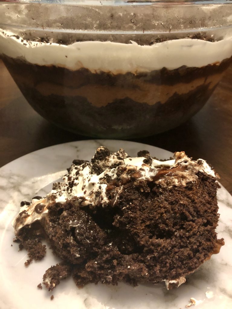 A serving of Chocolate Dirt Cake (Mud Bucket) on a white marbled plate. A trifle dish is behind the plate showing layers of chocolate and Cool Whip.