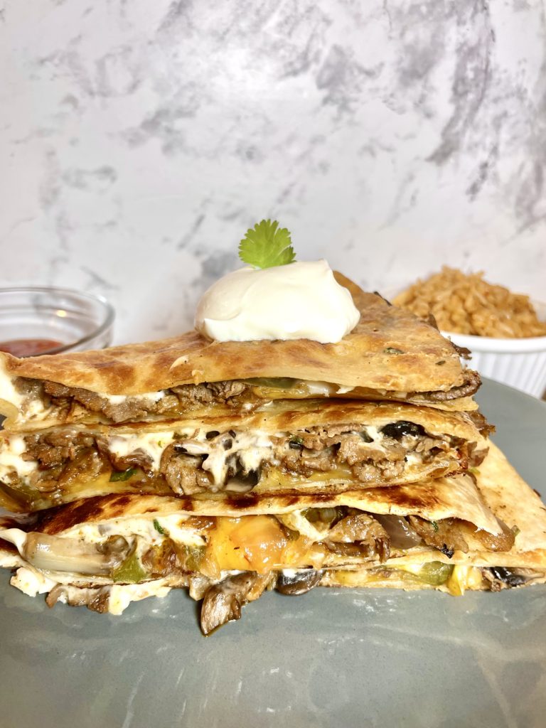 A stack of four steak bomb quesadilla pieces on a gray plate. A dollop of sour cream and a sprig of cilantro are on the top of the stack. There is a white ramekin in the background with Spanish rice and a red ramekin with salsa.