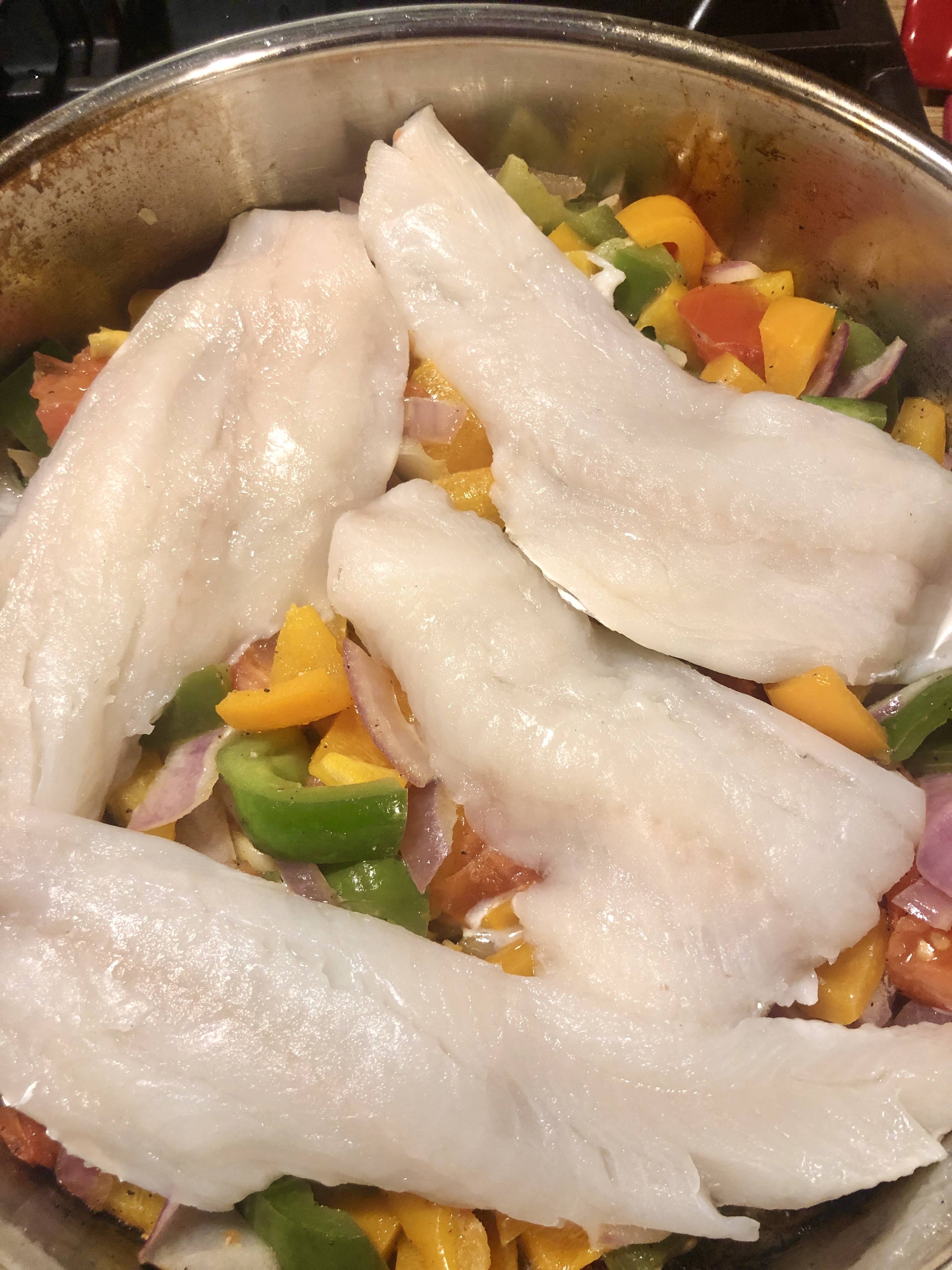 Four pieces of haddock in frying pan over cooking vegetables.