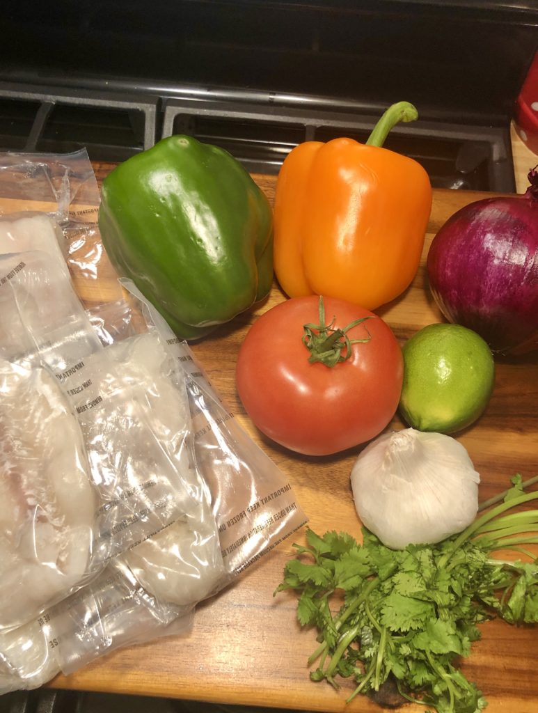 Ingredients for Moqueca (Brazilian Stew). Green and orange bell peppers, purple onion, tomato, lime, garlic, cilantro and haddock are pictured on a wooden cutting board.