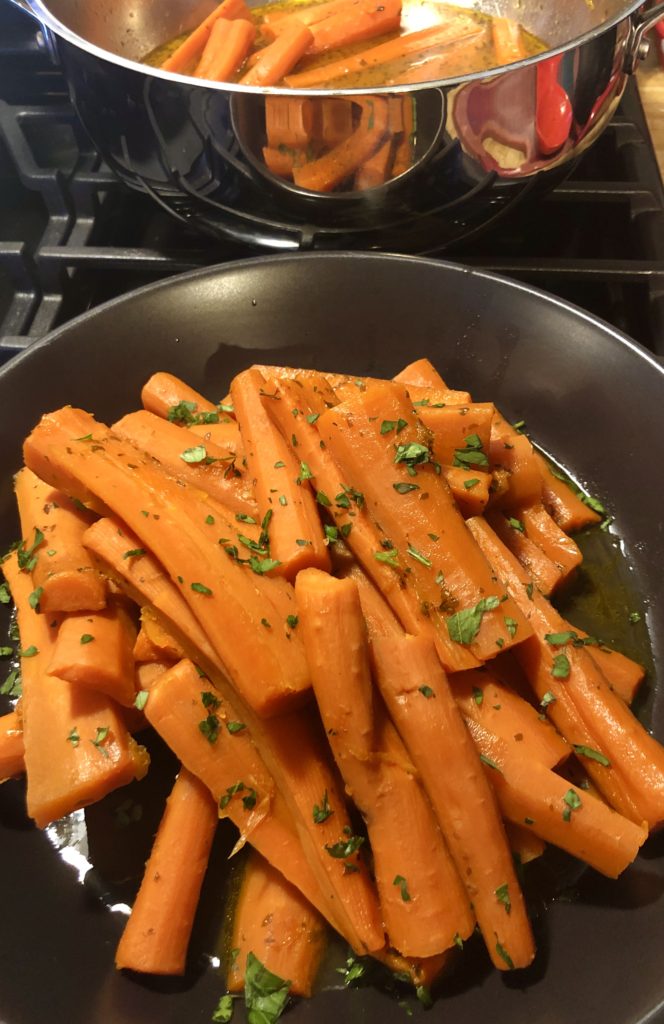Orange Apricot Glazed Carrots served in a black bowl and garnished with fresh parsley. There is a pan behind the bowl with additional carrots.