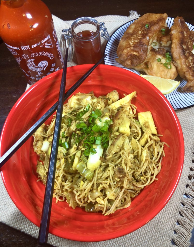 Singapore noodles in a red bowl with black chopsticks lying across the top of the bowl. Sriracha, a jar of chili sauce and a small plate of Chinese chicken fingers are behind the bowl.