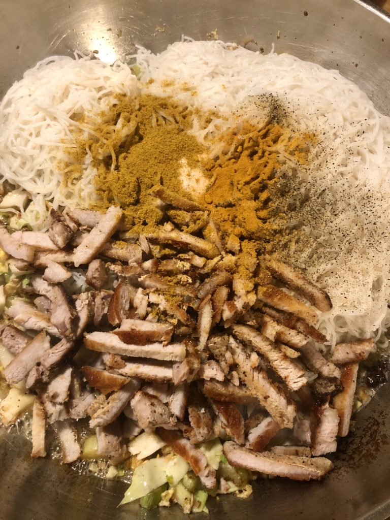 Curry powder and other spices added to frying pan with rice vermicelli noodles and cooked sliced pork.