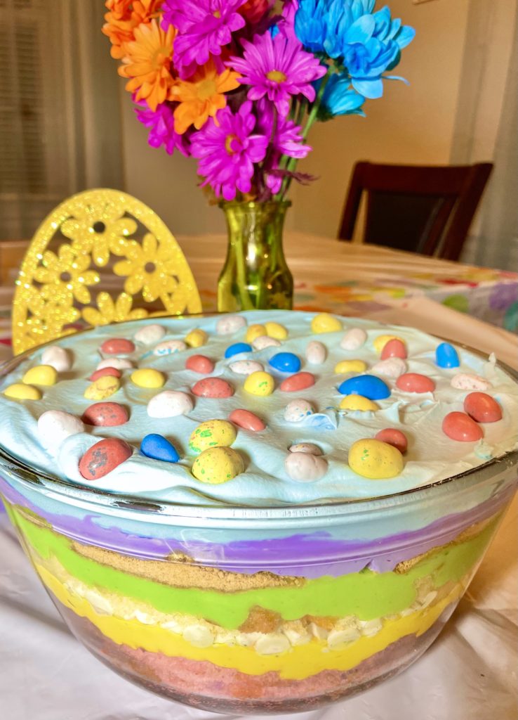The Best Easter Trifle Dessert pictured showing bright colorful layers of cake, vanilla pudding, funfetti morsels, cream cheese and Cool Whip. The trifle is topped with blue dyed Cool Whip and Whoppers Mini Robin Eggs.