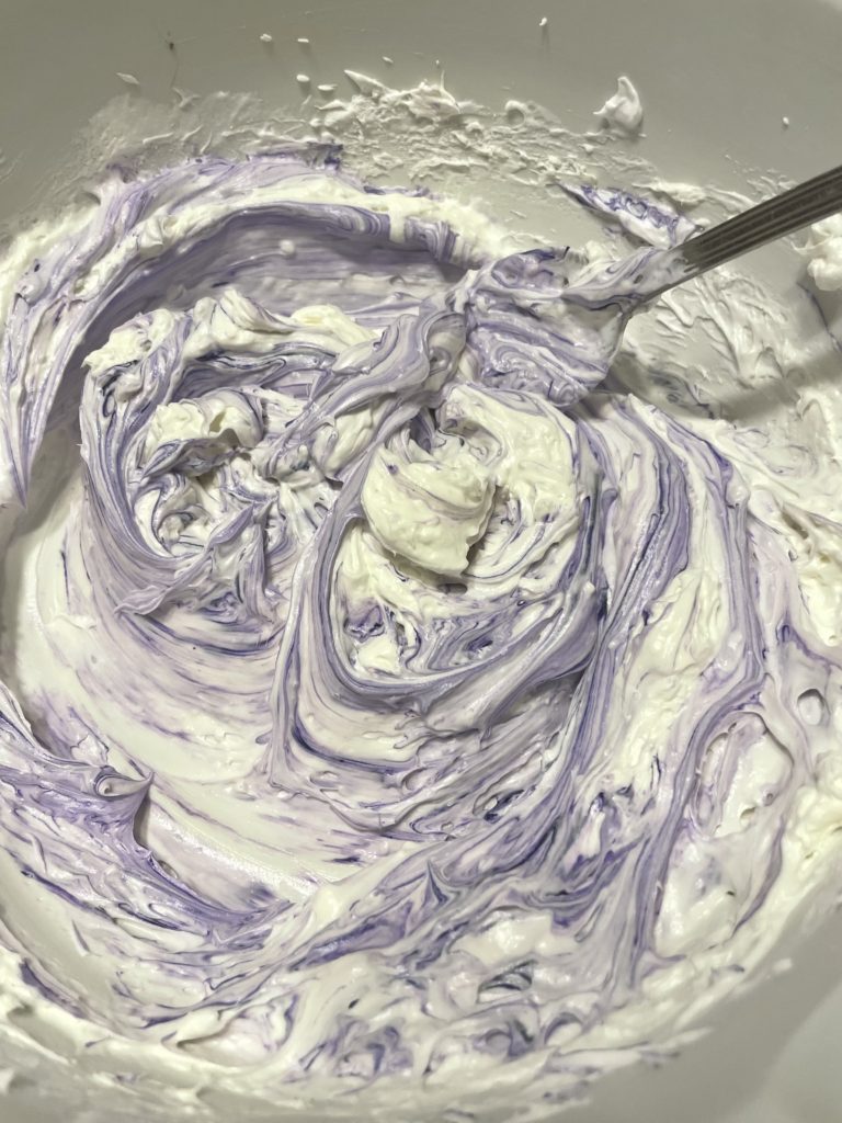 Whipped cream cheese and Cool whip in a mixing bowl with purple food color gel being mixed in.