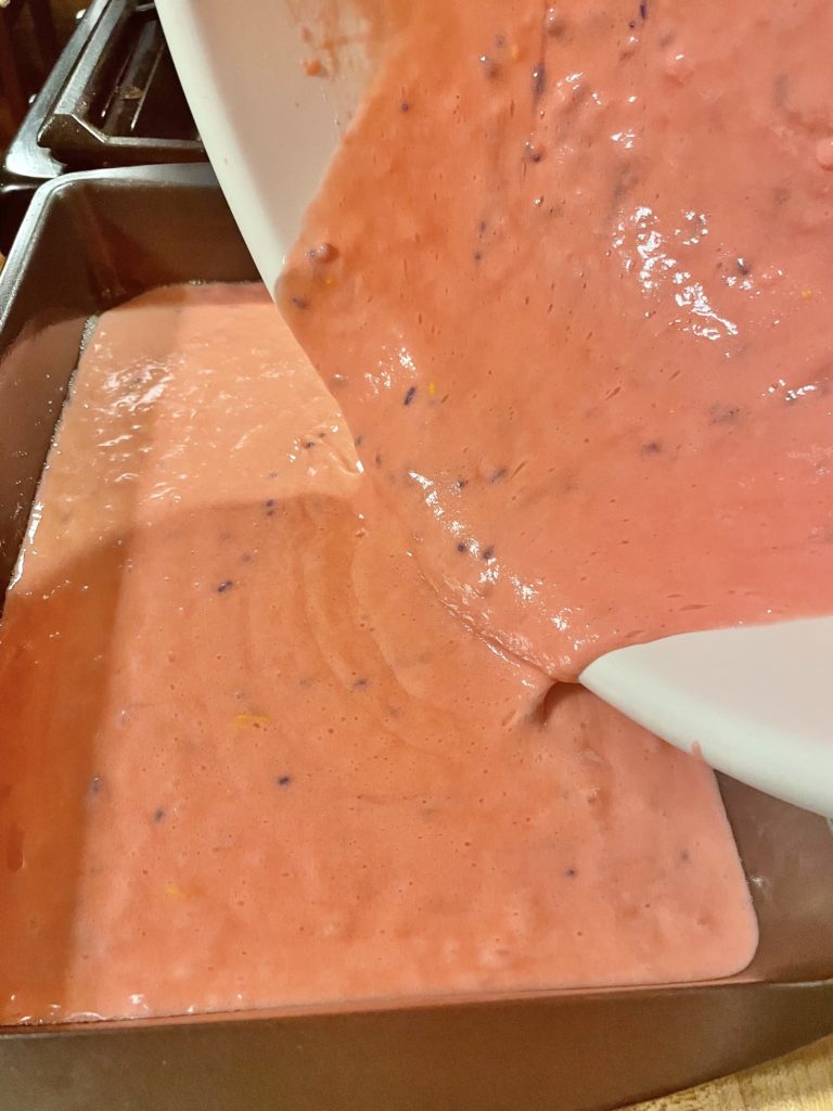 Prepared strawberry cake box mix being poured from white mixing bowl into 13x9 baking pan.
